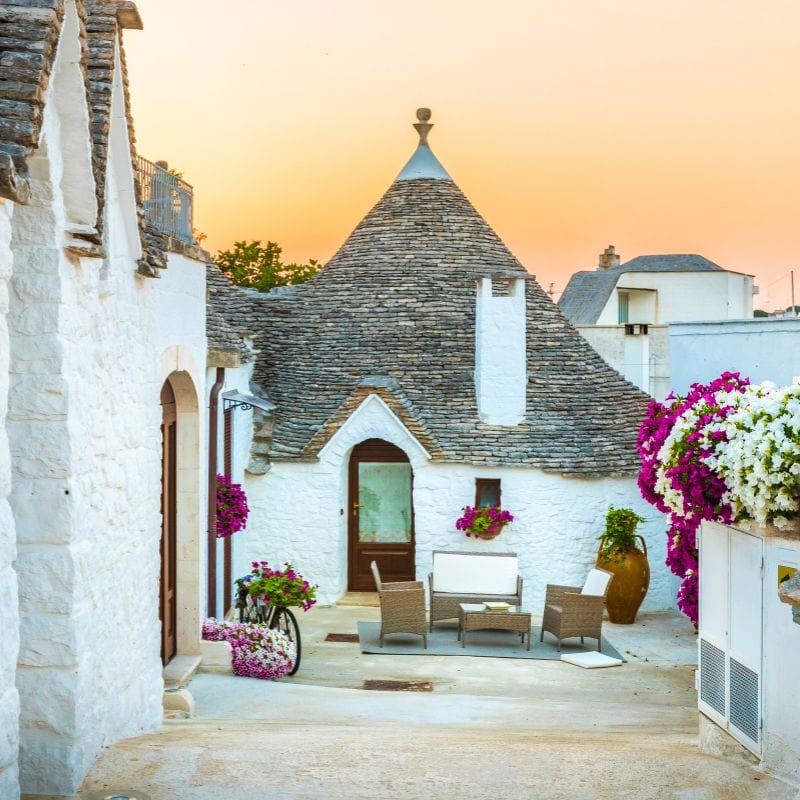 Alberobello image with purple flowers growing on white wall 