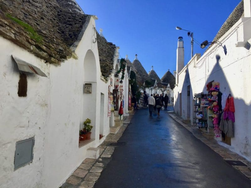 Alberobello street view - grey narrow lane with white fronted trulli shops either side. there are a few people in the street and some shops have goods hanging outside 