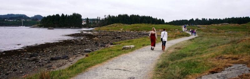 Two women walking across the homeland of the vikings towards the viking farm. Landscape is flat beside path with rough grass .In the distance is the island with pine trees.
