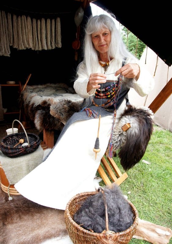 Woman dressed as a viking with a white dress and a blue cloak over top. She has long white hair and holding a woodenspindle and spinning grey wool into yarn