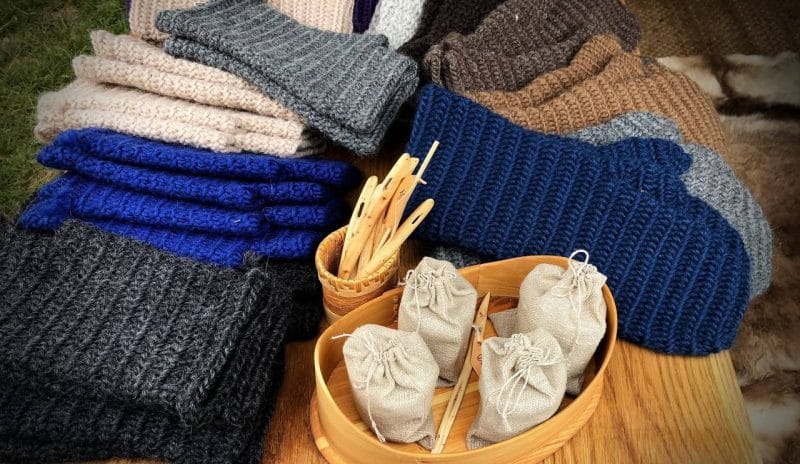 Blue, grey and brown mittens, hats and neck warmers displayed on a table with a pot of wooden large needles.