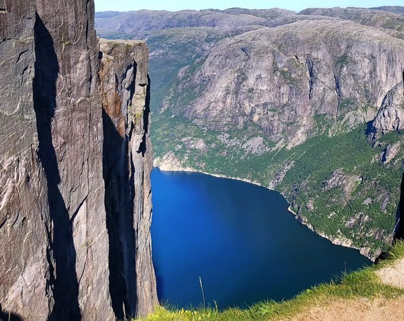 A view down into the deep blue fjord below. On the left a grey brown rock-face and around the fjord tall granite mountain with patchy green