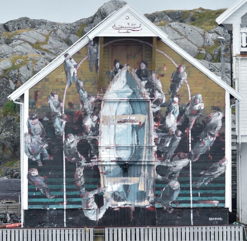 A large mural on the side of a house with a fishing boat in the middle and people stood around it. It is painted from a birdseye view and the colours are mainly pale grey, dark grey brown and blue.