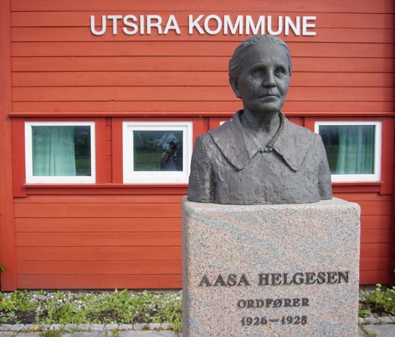 The grey bust of the first mayor of the island, Aasa Helgesen in front of the red wooden building of the Utsira municipality