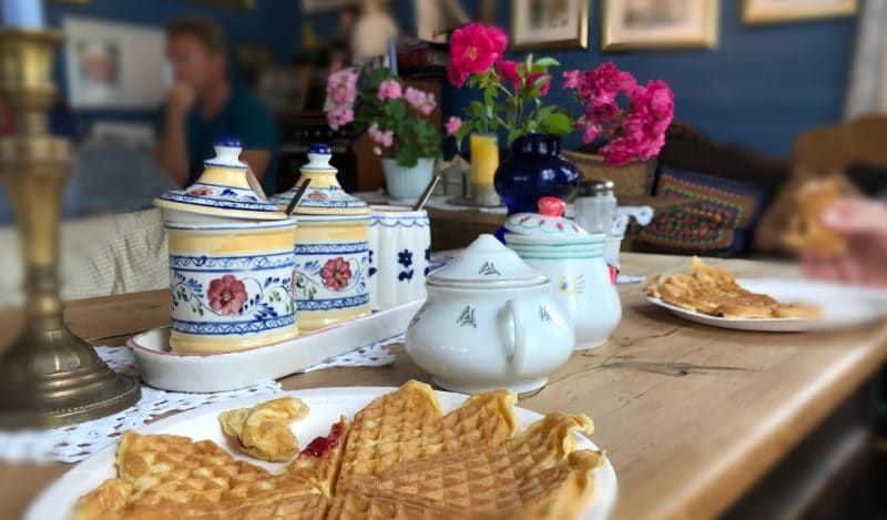 A white plates has a waffle on it that has a slice taken out of. The background is blurry with little yellow, blue white and red pots in the background.