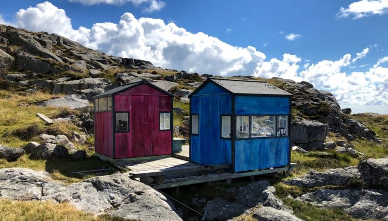 Utsira Huts :Two smal huts, one pink, one blue on the craggy landscape of rocks and grass 