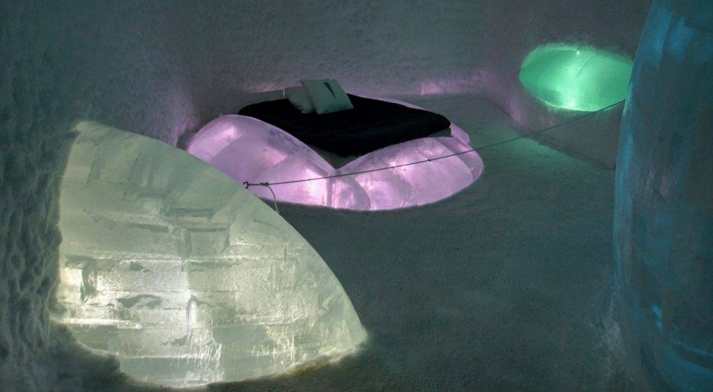 The bed in the middle of the photo is on a pinkish block of ice, there is a green backlit ice shelf to the left and a white half igloo on the left