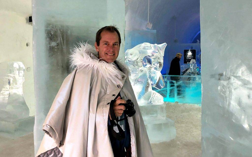 Lars, wearing a stone coloured lined thick cape with furry collar. The ice sculptures in the background. 
