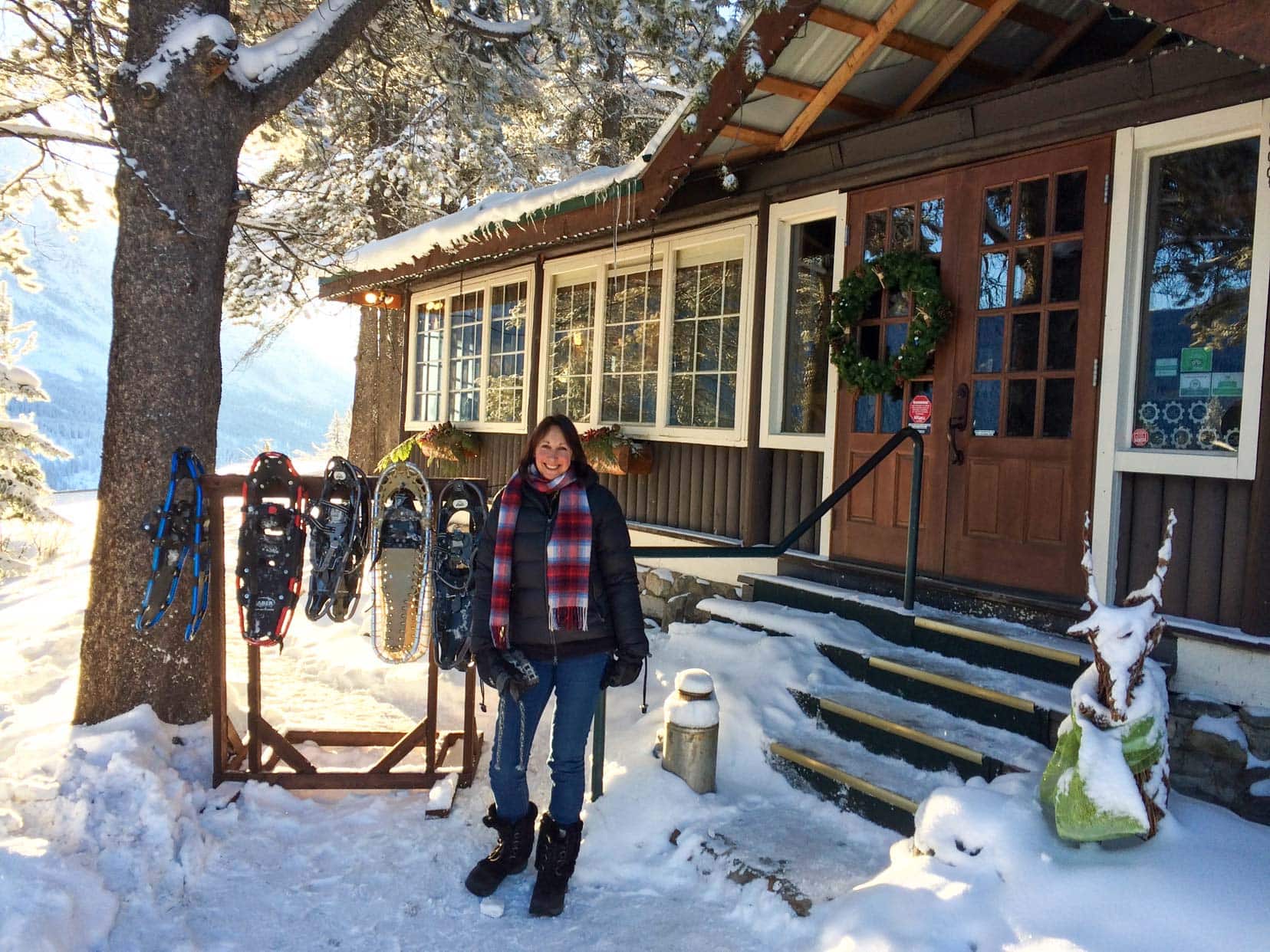 Shelley stood by a log cabin in the snow by a rail of snowshoes hanging by a tree 