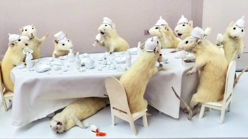 An art instalation at the National Art Gallery of Tirana, Albania. 10 stuffed white dwarf hamsters in poses around a white table that is sized relative to them. 