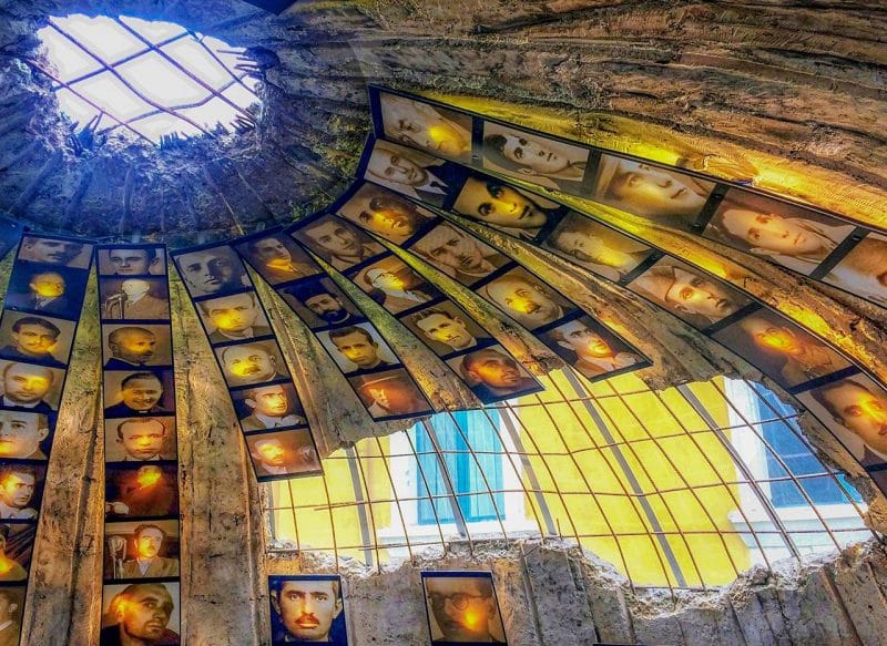 A dome shaped bunker with photographs of men arranged vertically around the dome. There is damage to the outside, so you can see through to the yellow wall of the building outside.