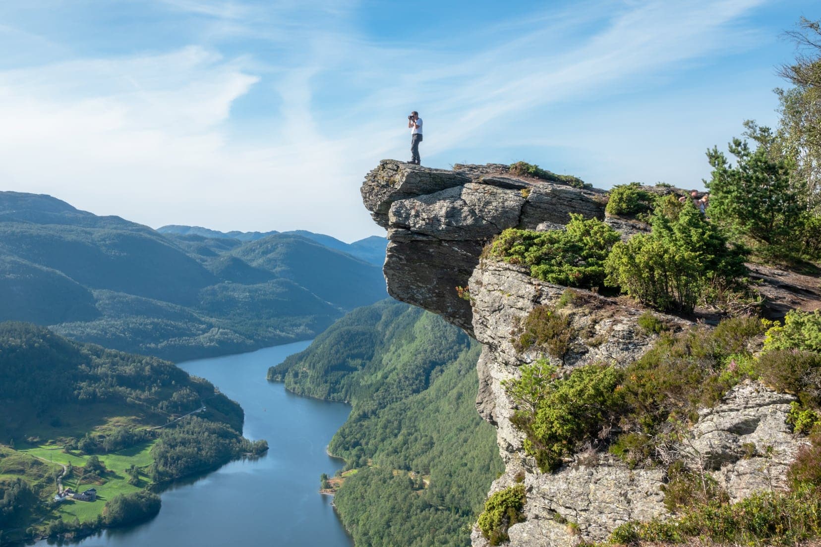 Man taking photograph from top of rocky outcrop with a fjord below