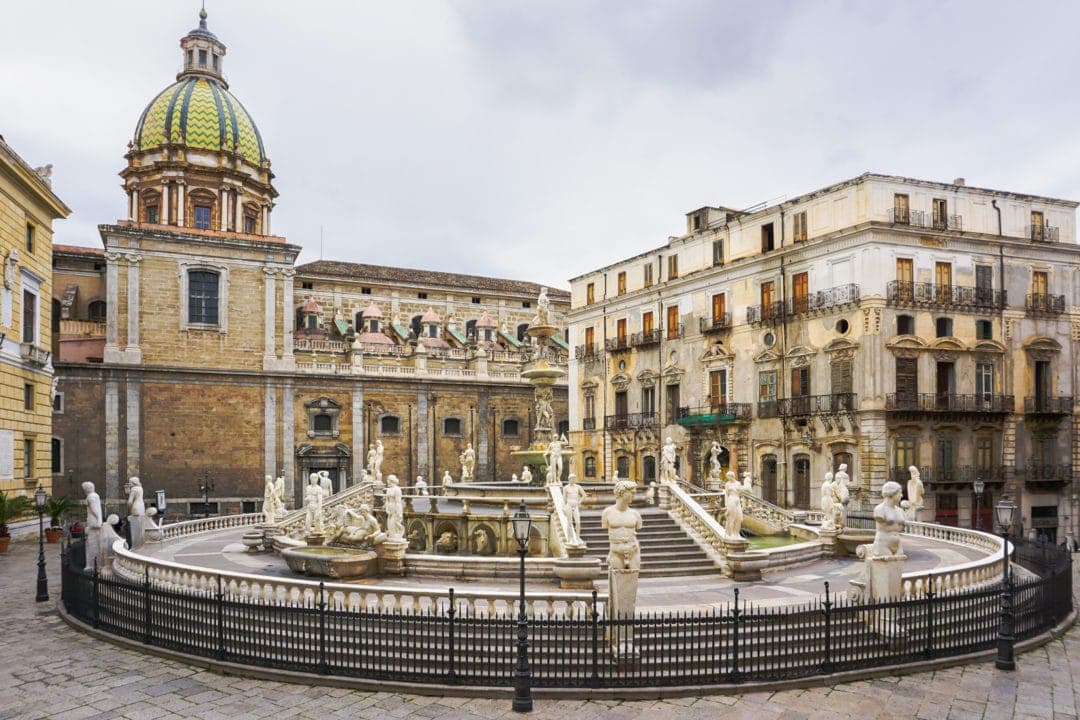 Fountain with many statues around surrounded by buildings of marble