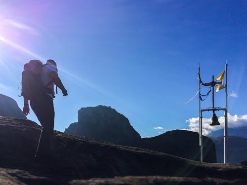 Man with backpack walking towards a cross and bell on a mountain top