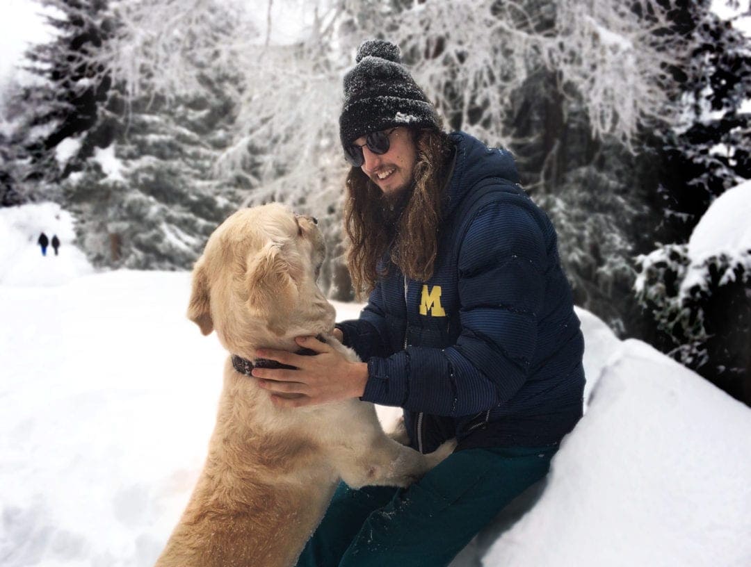 young man with long hair dressed in blue winter gear in the snow patting a golden retriever and smiling