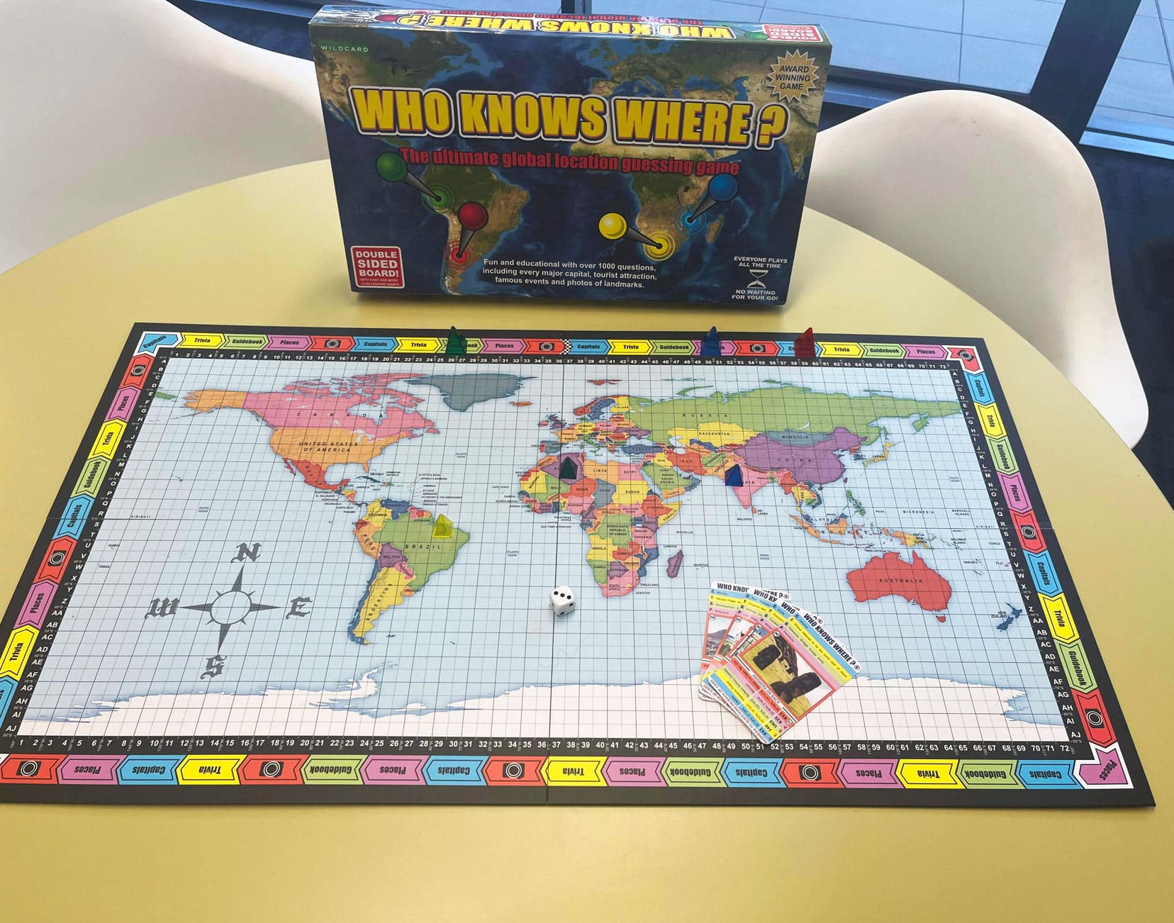 Our Who Knows Where game playing board with some cards on a world map with a dice and two small cubes