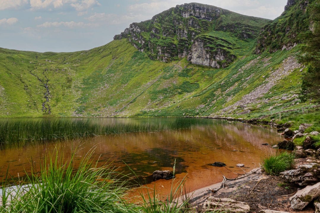 Brown coloured lake water is surrounded by mountainous grassy slopes