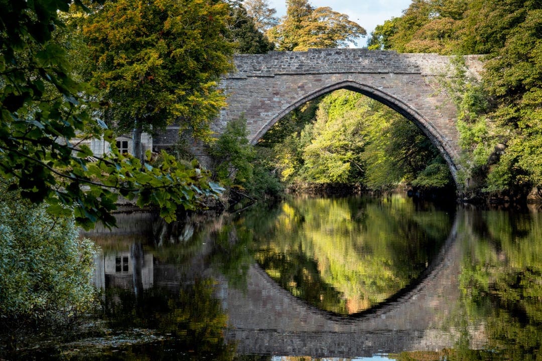 the bridge with a reflection of its arch in the water and green trees either sidef
