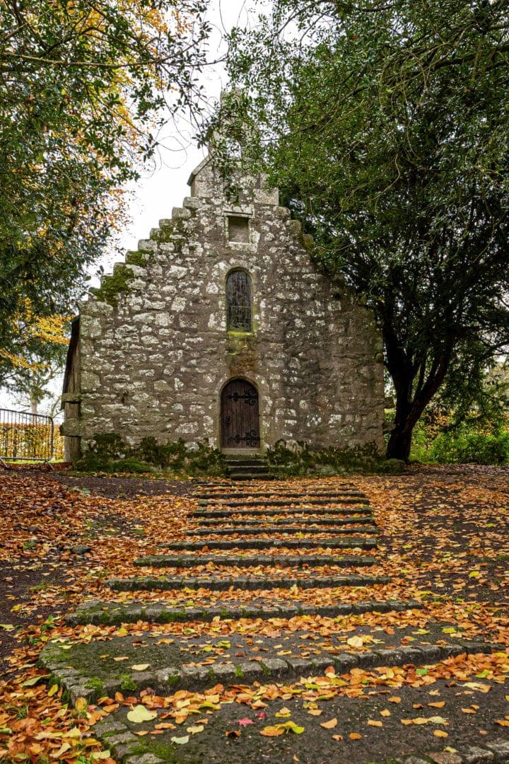 A triangular roofed grey stone chapel with steps covered in autumn leaves leading up to the entrance 