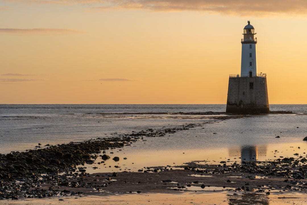 Rattray Head lighthouse with orange hued sky and low tide showing rocks and sand in the foreground