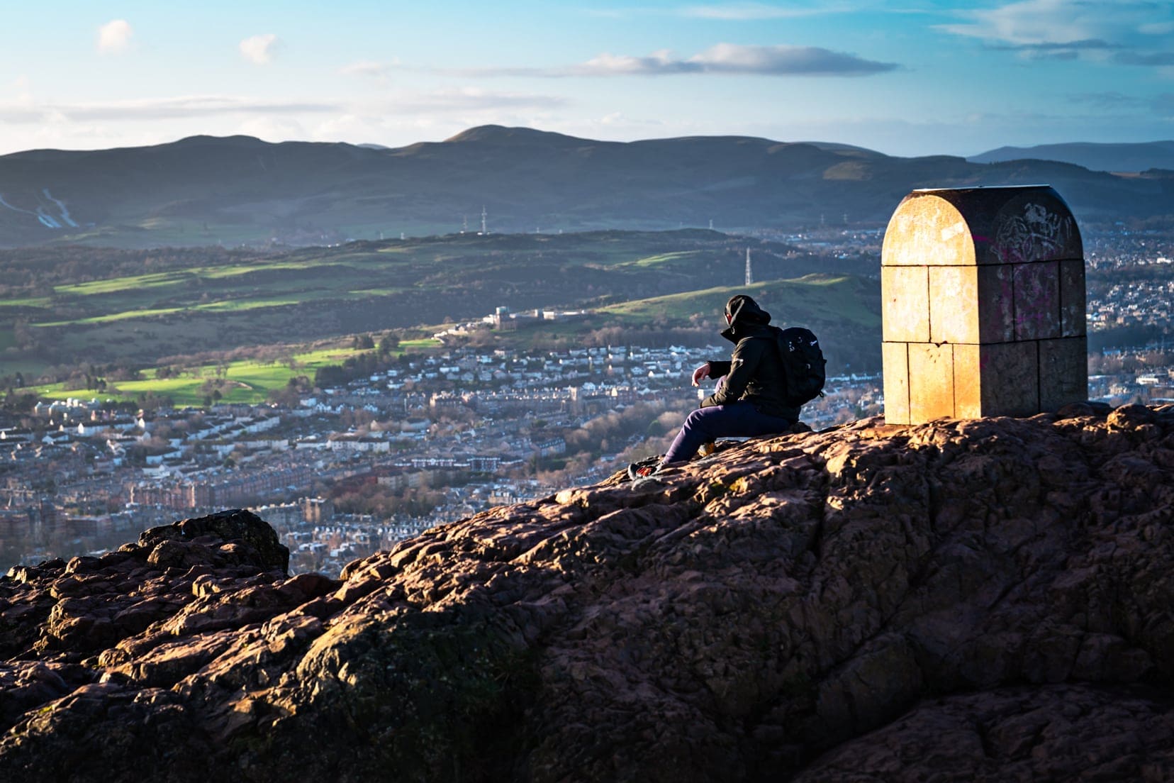 Man sat beside the concrete block on top of Arthur's Seat in Edinburgh looking out to view beyond