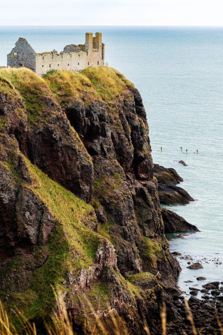 Portrait-of-dunnottar-castle showing the steep cliff face to the south and the tiny figures of paddleboarders on the ocean beside it