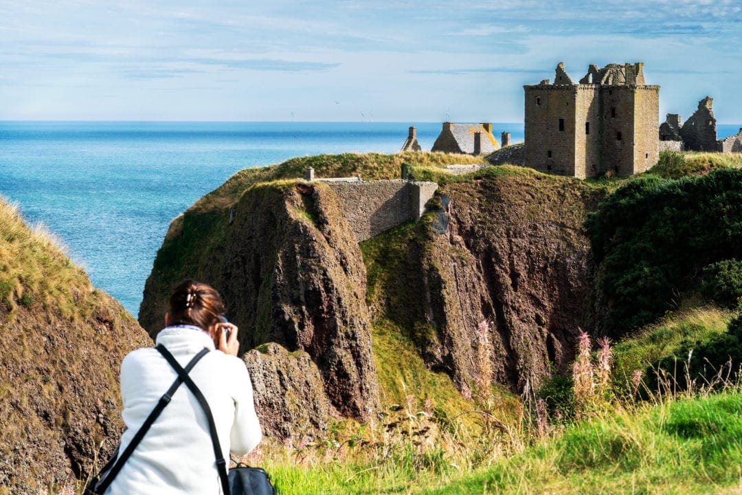 Woman bent down taking a photo of Dunnottar castle in the distance