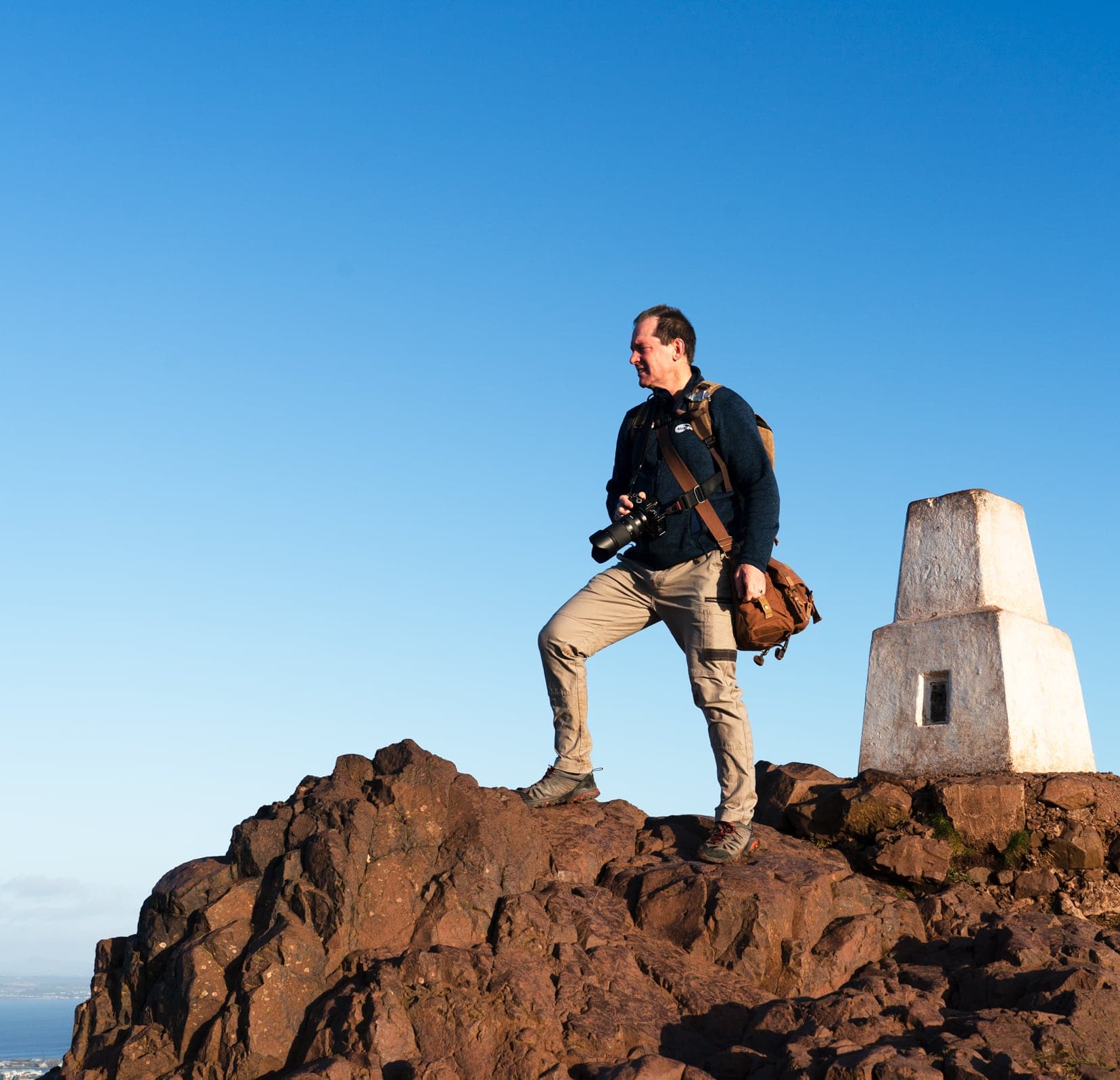 Lars stood by a white two layerd concrete block on top of brown craggy rocks at the summit of Arthur's Seat
