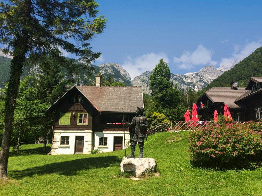 Alpine hut with a statue in front in a green meadow wiith mountains in the background