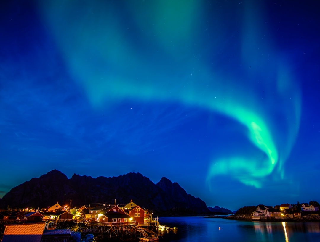 A swirl of green lights - the northern lights over a distant mountain and the village of Henningsvaer lofoten