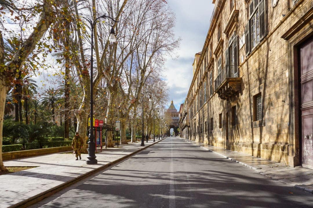 Long street in Palermo with trees lining one side and terrace buildings on the other. At the end is an old arched gateway