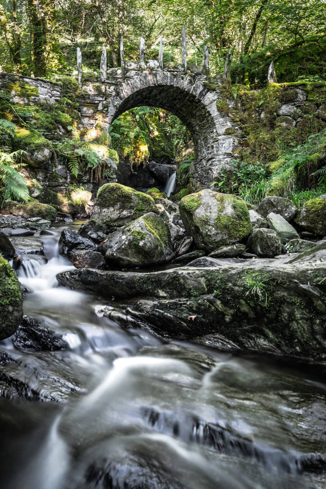 arched stone bridge with a waterfall behind and water blurred through long exposure