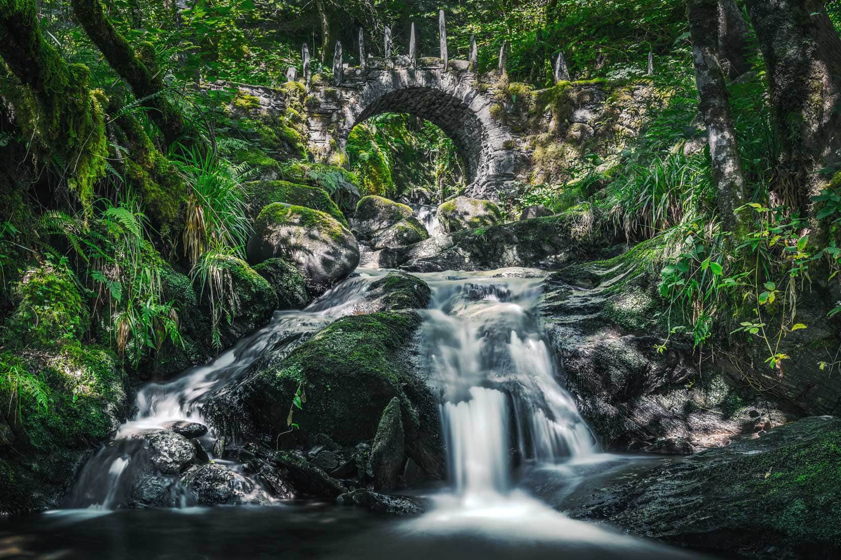 long exposure photo of a stream under an arched stone bridge with vertitical rocks