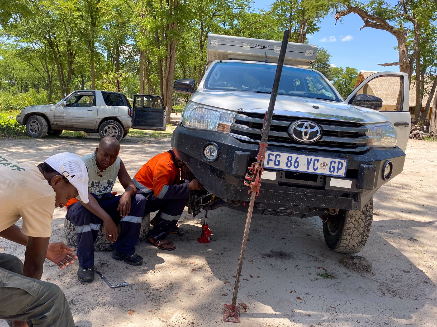 two mechanics with our Hilux jacked up with the front front wheel off in a campsite within Moremi national park, Botswana