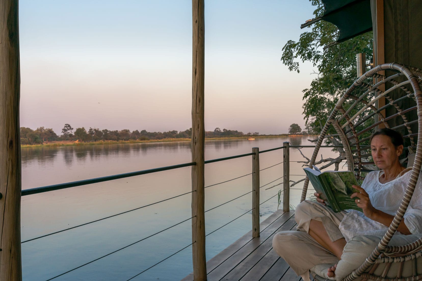 Shelley sitting on a chair reading a book on the deck with the Okavango River beside her