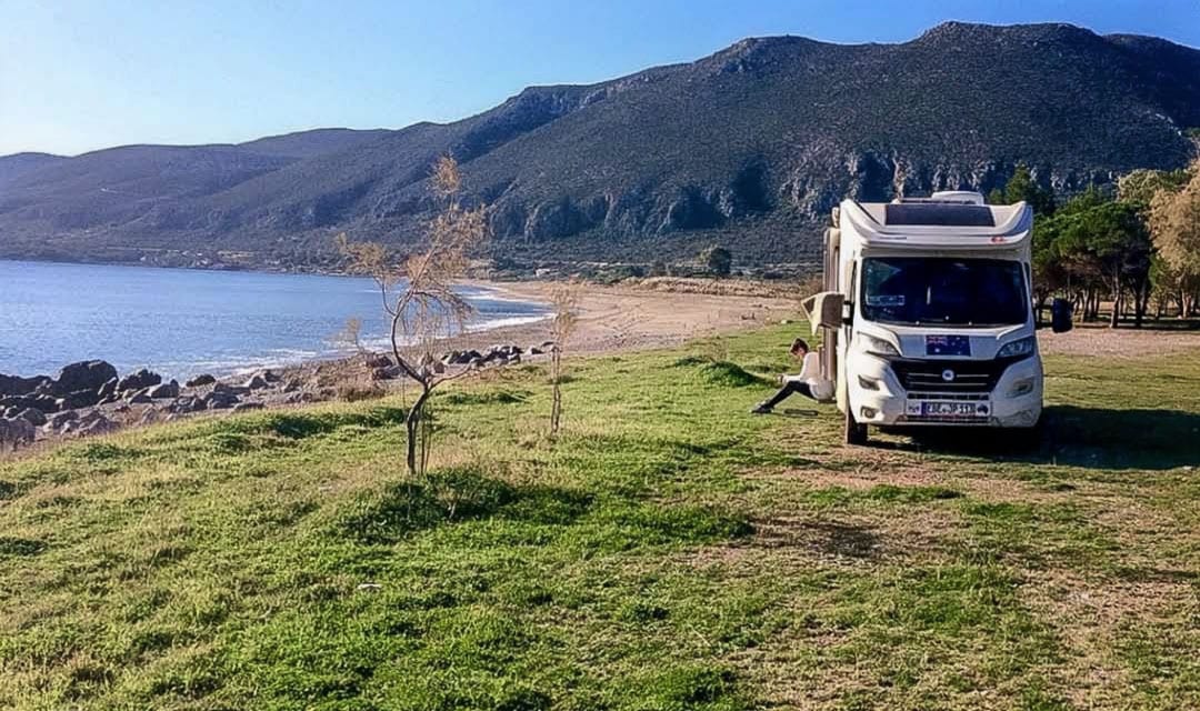 Shelley sat on the steps of the motorhome which is parked by the ocean on some grass with mountains in the background