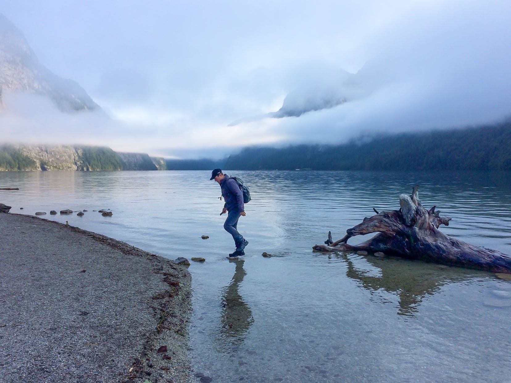 Lars jumping on stepping stones om a river away from an old log with mountains, trees and fog in the distance.