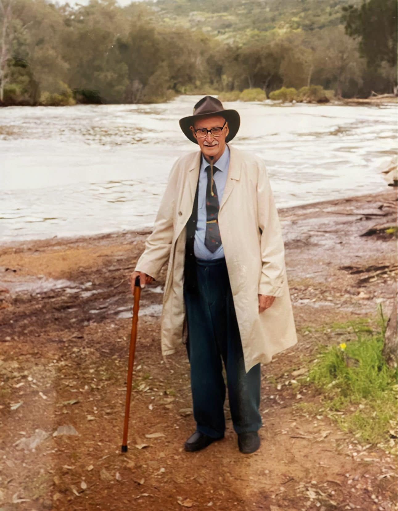 my grandad stood by a river with trees in the back ground in WA. He is wearing a shirt and tie ans raincaot and holding a walking stick 