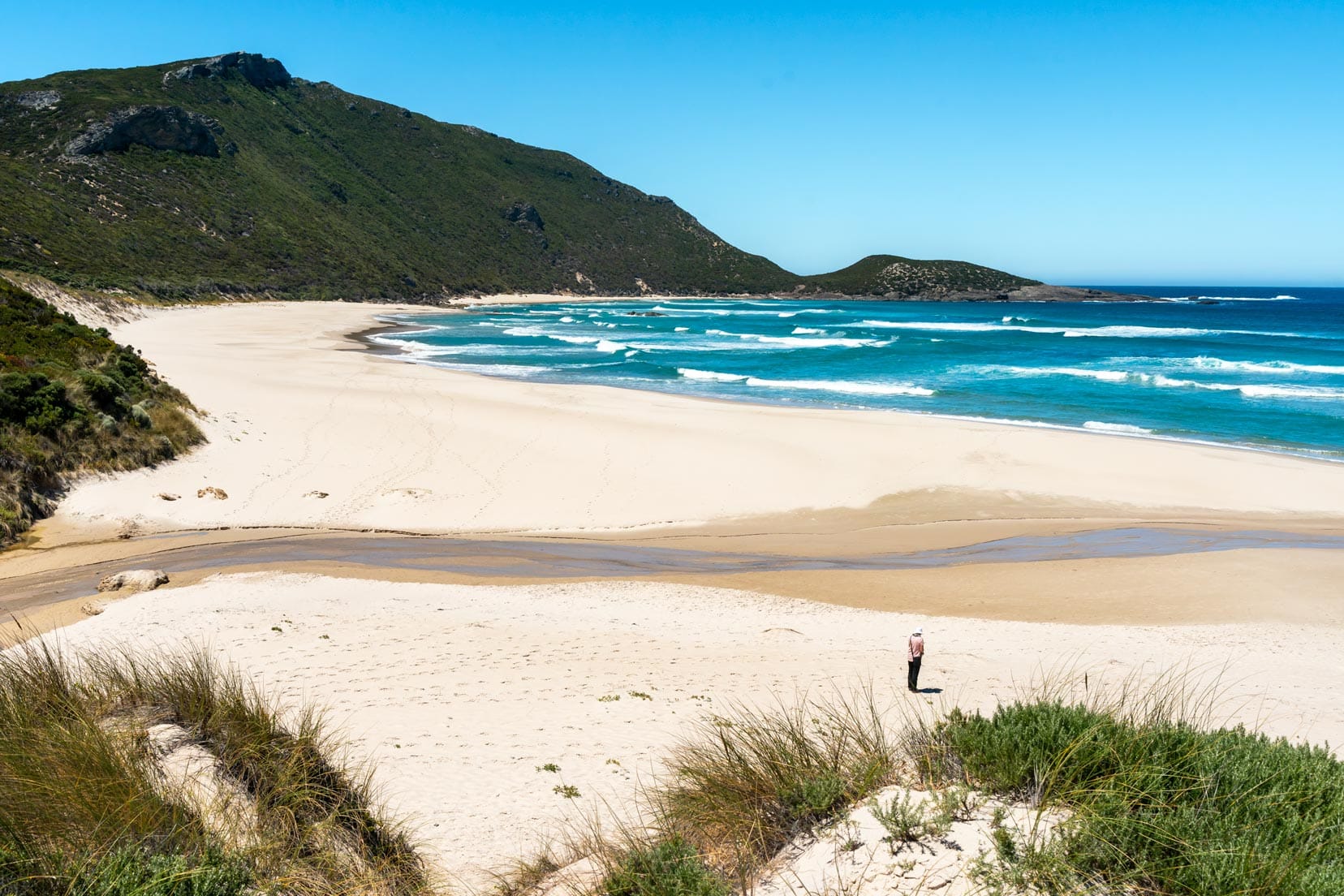 Shelley stood on Conspicuous cliff beach with a sweeping bay of white sand and a backdrop of green hills 