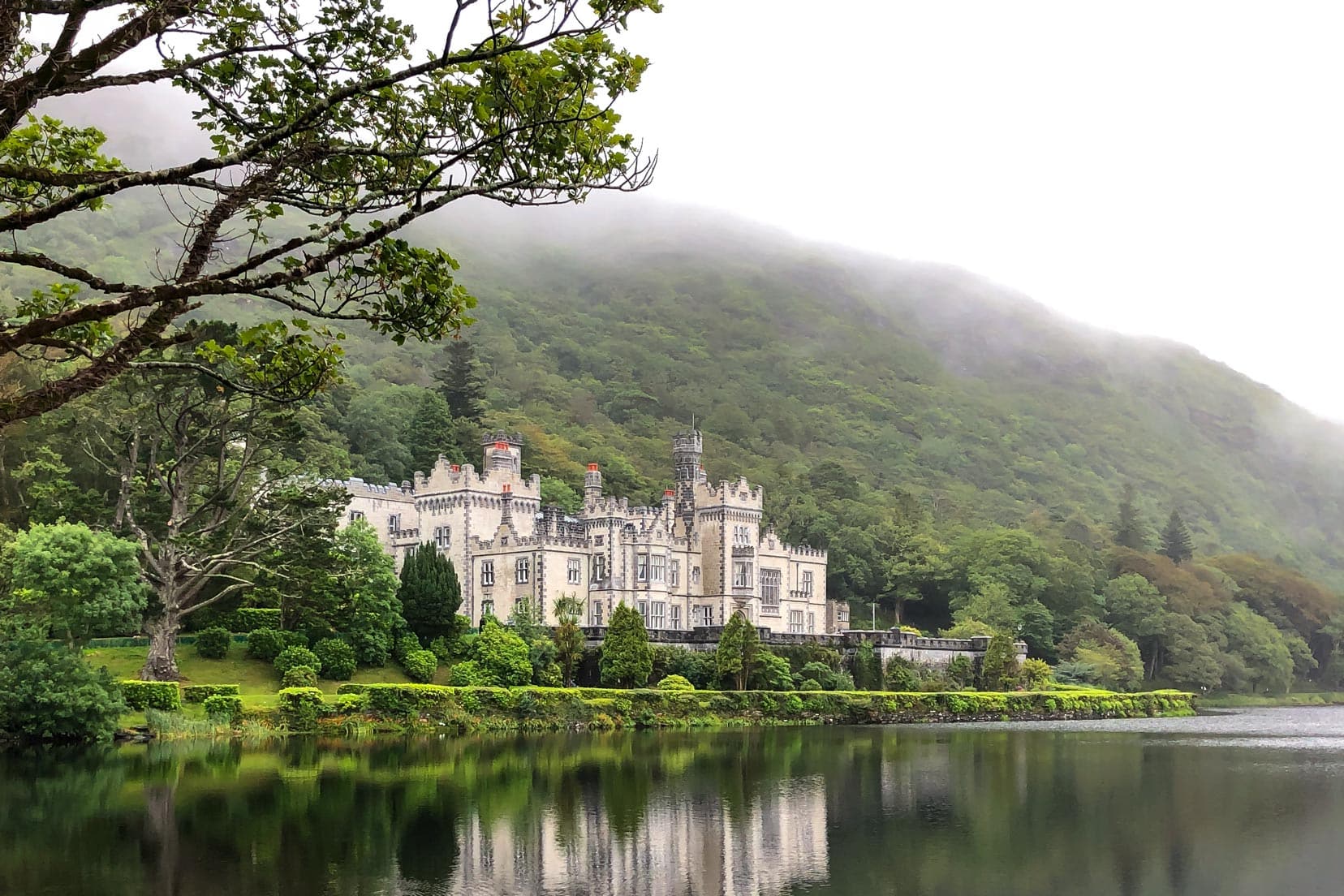 Kylemore-Abbey-seen-from-path-leading-in with a lake in the foreground and forested hill in the background