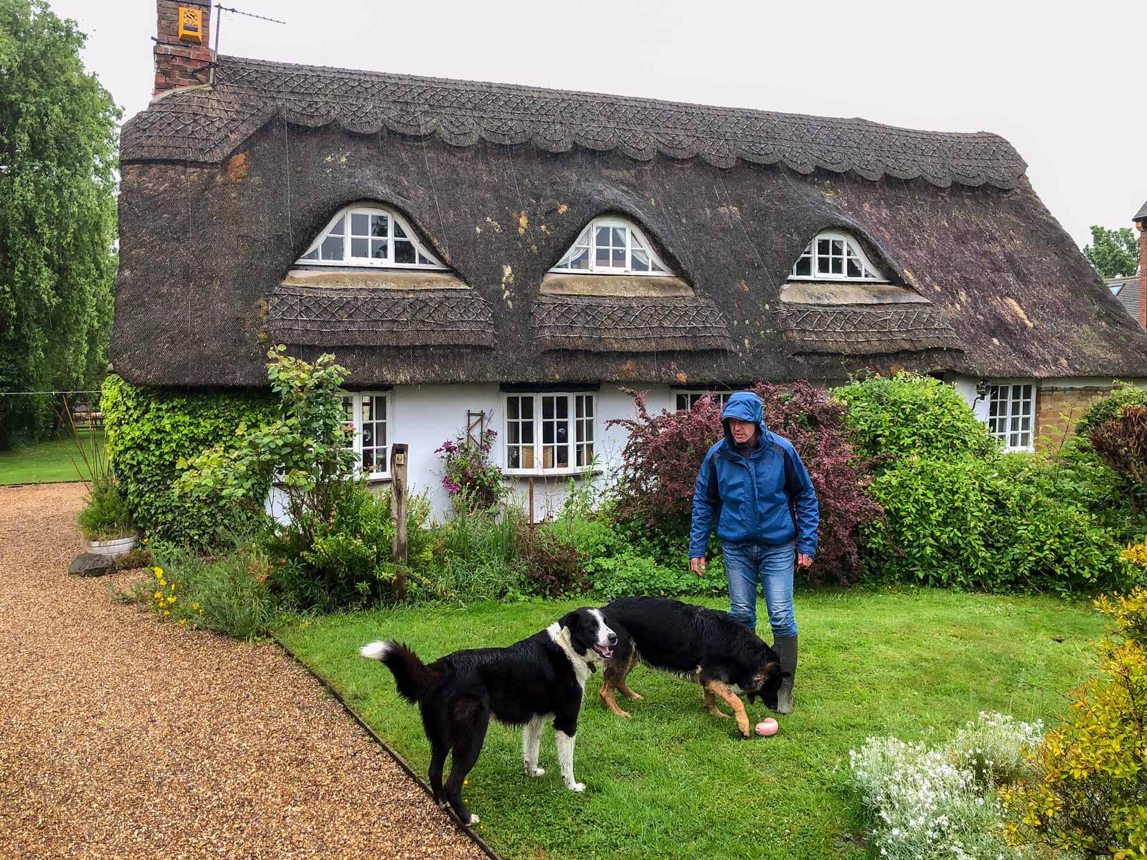 Exploring the cotswolds on a house sit in the uK - Lars stood in a graden with two dogs with the house we stayed in in the background with a beautiful shaped thatched roof