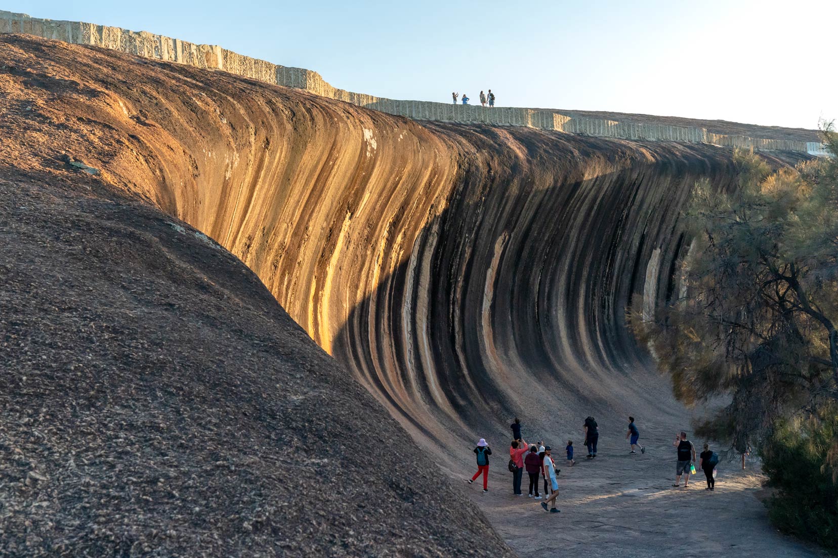 Wave rock shaped like a wave with sun hitting part of it and a few people at its base