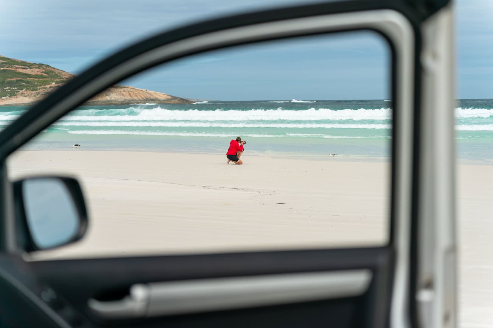 Beach-car framing - Shelley kneeling on the sand by the ocean and can be seen through the opened window of the car door 