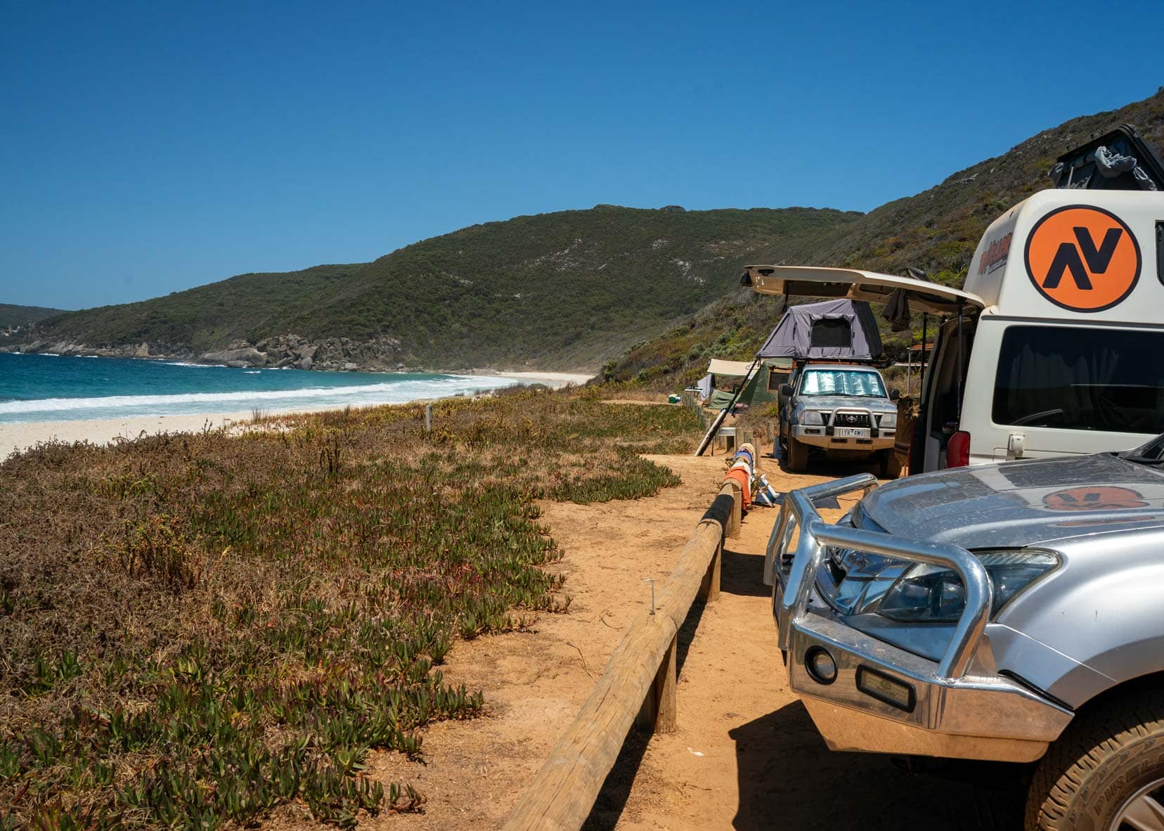 Shelley-Beach,-Cape-Howe cars parked on sandy area by beach with green hills and ocean in background